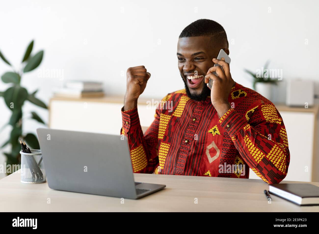Un homme d'affaires africain débord de joie dans des vêtements traditionnels célébrant le succès sur le lieu de travail Au bureau Banque D'Images
