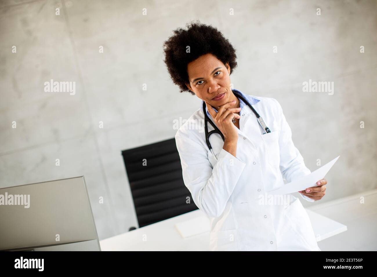 Femme afro-américaine médecin portant un manteau blanc avec stéthoscope debout par bureau dans le bureau et en regardant les tests médicaux résultats sur papier Banque D'Images