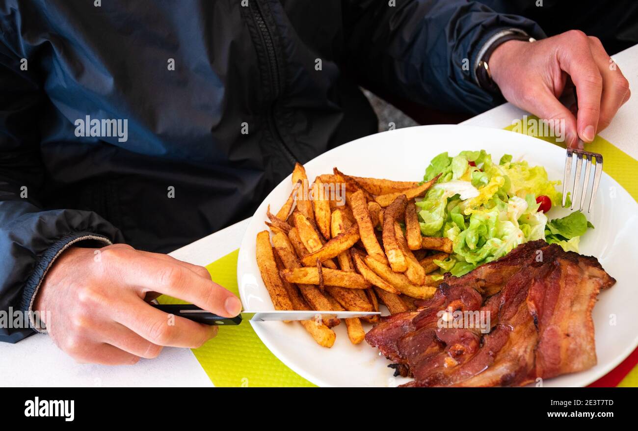 Un homme qui travaille dur pour un déjeuner nutritif. Poitrine de porc grillée, frites et salade verte. Banque D'Images