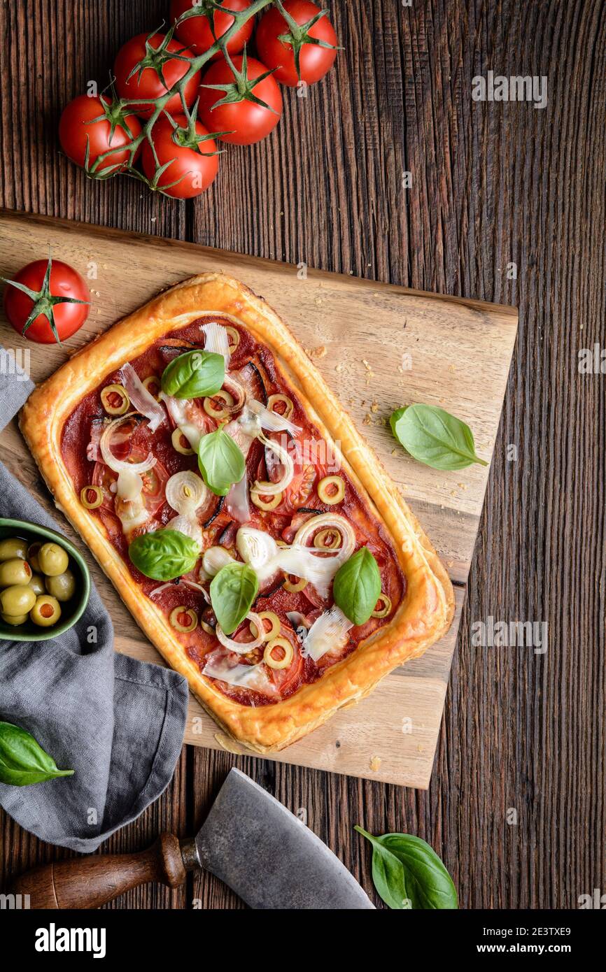 Pizza feuilletée maison avec tranches de bacon, tomates, olives vertes, fromage et oignons, recouvertes de feuilles de basilic sur fond rustique en bois Banque D'Images