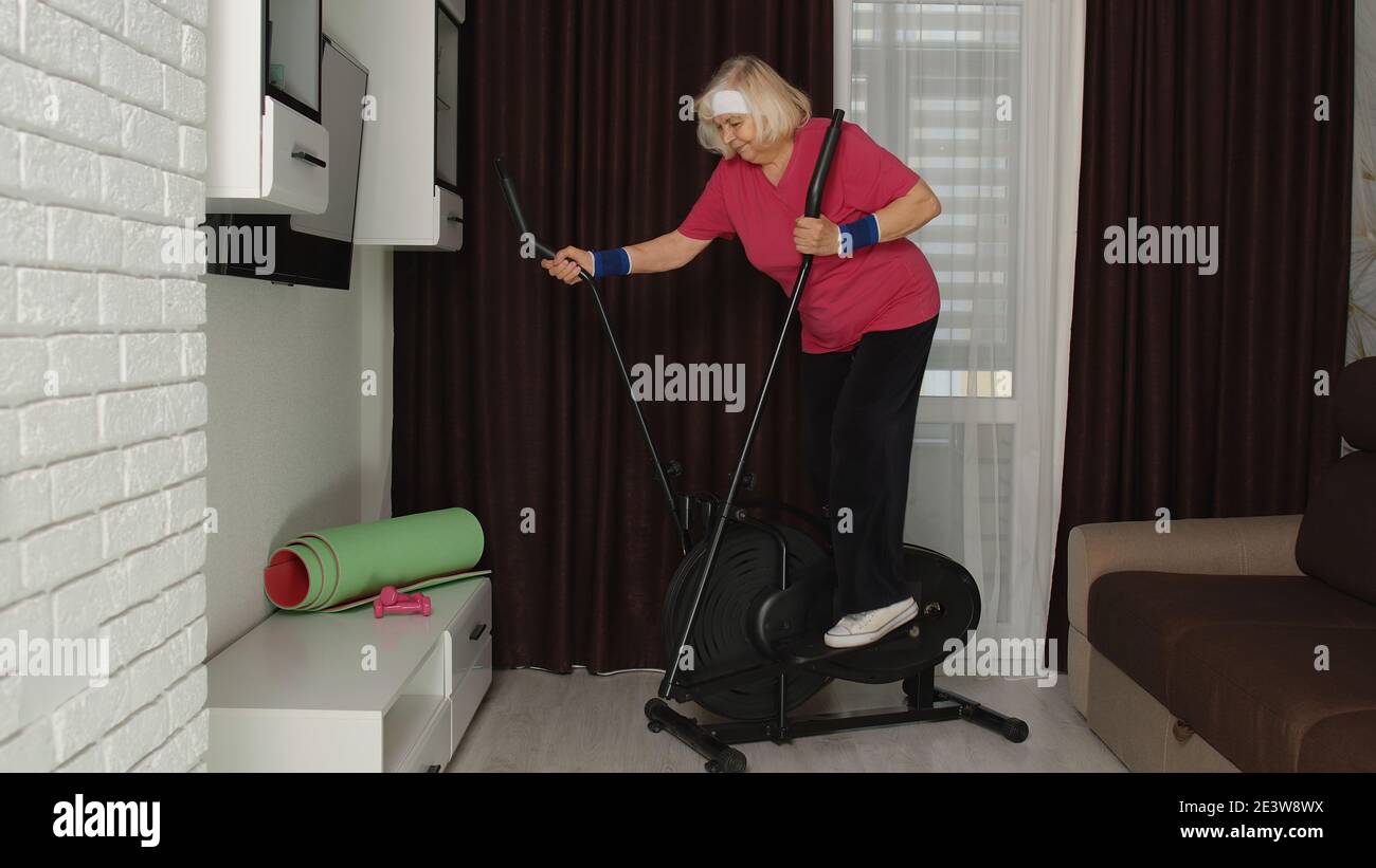 Ancien modèle de grand-mère sénior aux cheveux gris faisant de l'exercice sur orbitek dans la chambre à la maison. Style rétro caucasien mature femme âgée s'échauffe tout en s'asseyant sur orbitrek intérieur. Un mode de vie sportif sain Banque D'Images