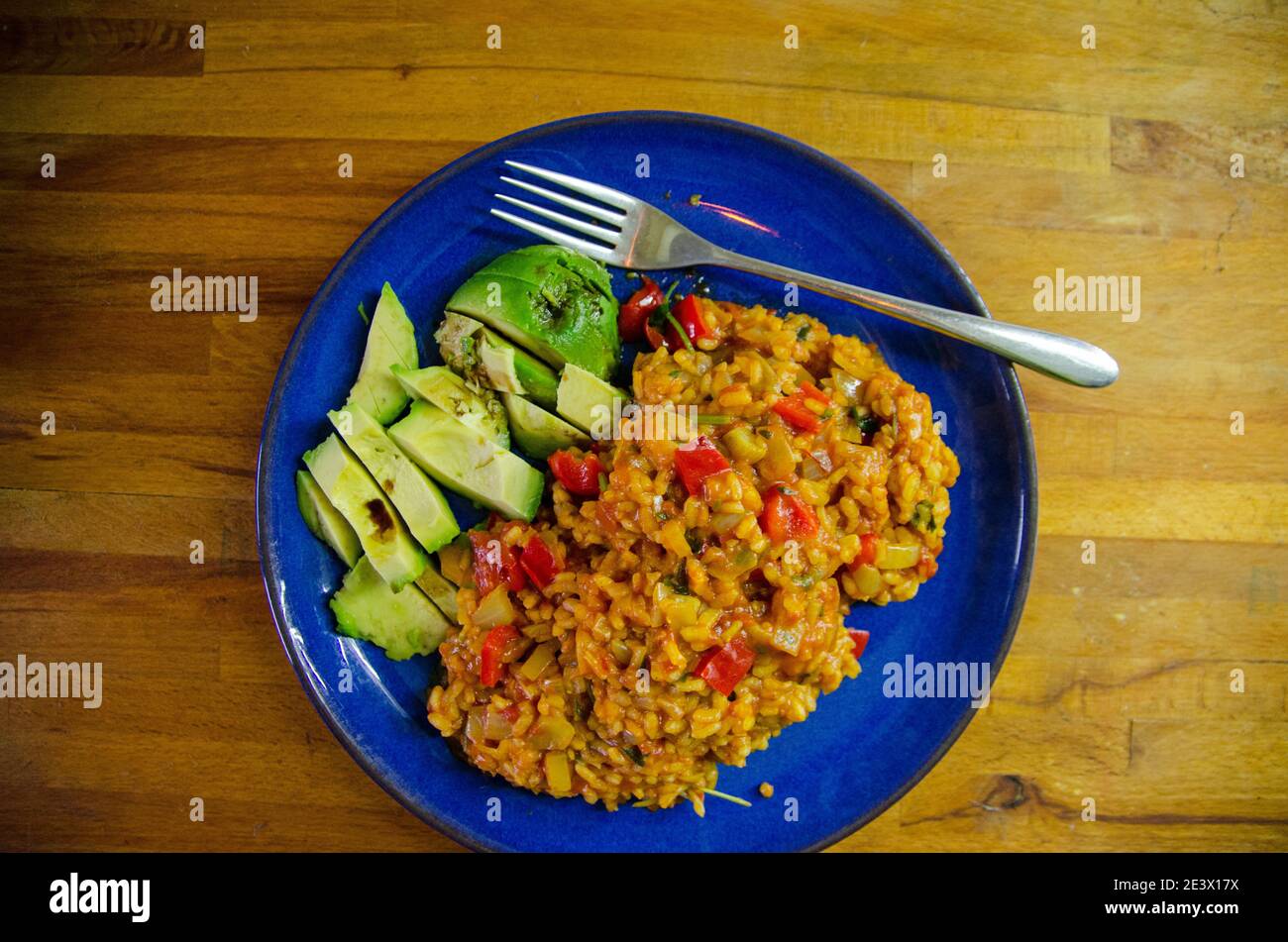 Vue aérienne d'une assiette bleue contenant de la paella végétale servie avec une tranche d'avocat frais vêtu de vinaigre balsamique. Banque D'Images