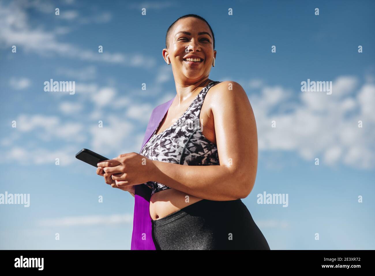 Femme en bonne santé dans les vêtements de sport avec téléphone mobile après la séance d'entraînement. Femme aux courbes qui prend une pause après l'entraînement en regardant loin et en souriant à l'extérieur. Banque D'Images