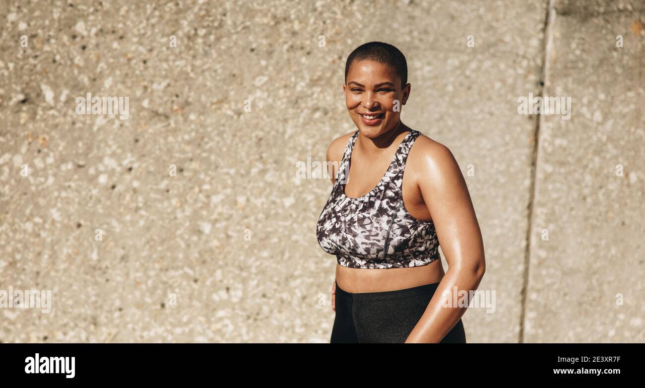 Portrait d'une femme heureuse en vêtements de sport debout près d'un mur à l'extérieur et souriant. Femme avec une coiffure à la mode se détendant après l'entraînement. Banque D'Images