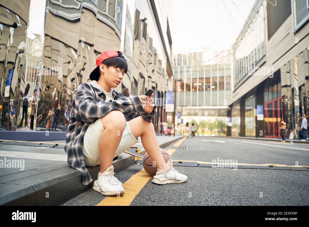 adolescent asiatique avec basketball et planche à roulettes assis sur le trottoir de la rue donnant sur le téléphone portable Banque D'Images