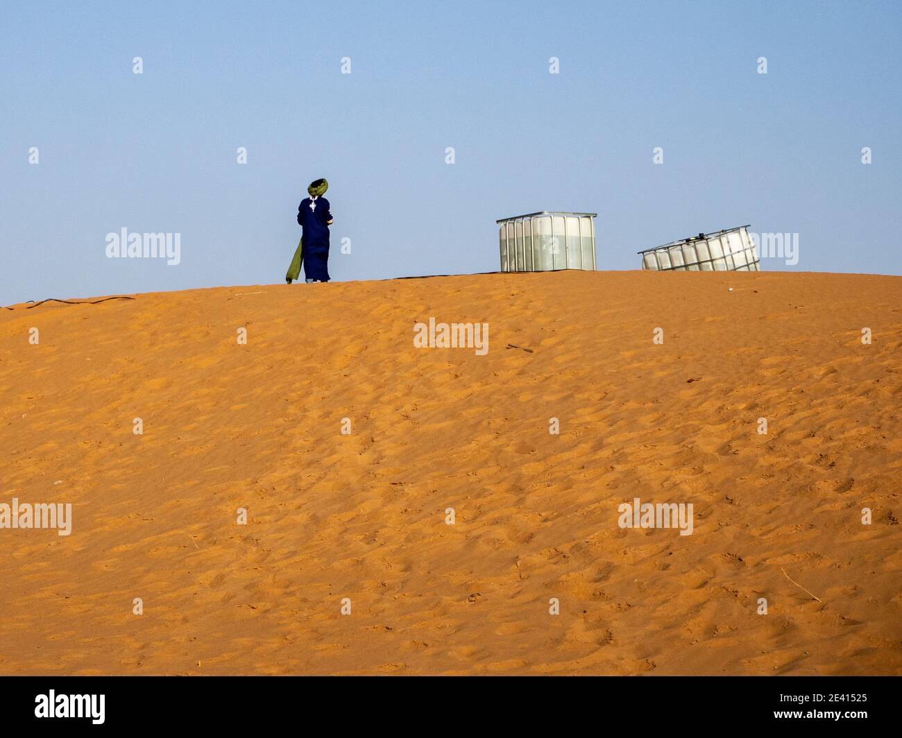 Un homme en robe berbère bleue typique avec turban, à côté de deux réservoirs d'eau en plastique sur une grande dune de sable dans le désert du Sahara marocain. Banque D'Images