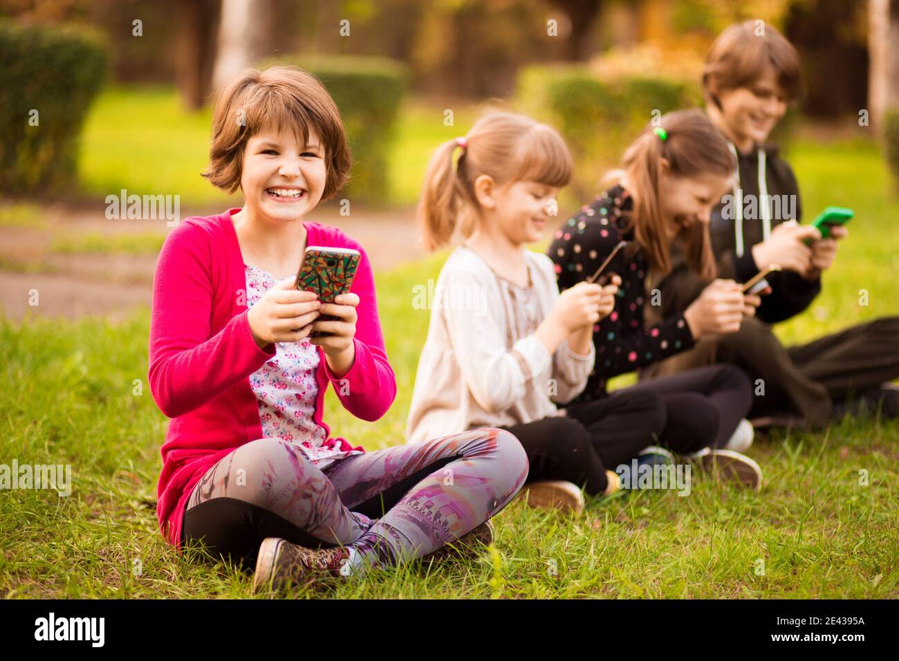 Photo pleine longueur d'un portrait extérieur d'enfant positif inspiré, fille assise sur l'herbe avec les jambes croisées et en utilisant le téléphone portable en compagnie d'amis Banque D'Images
