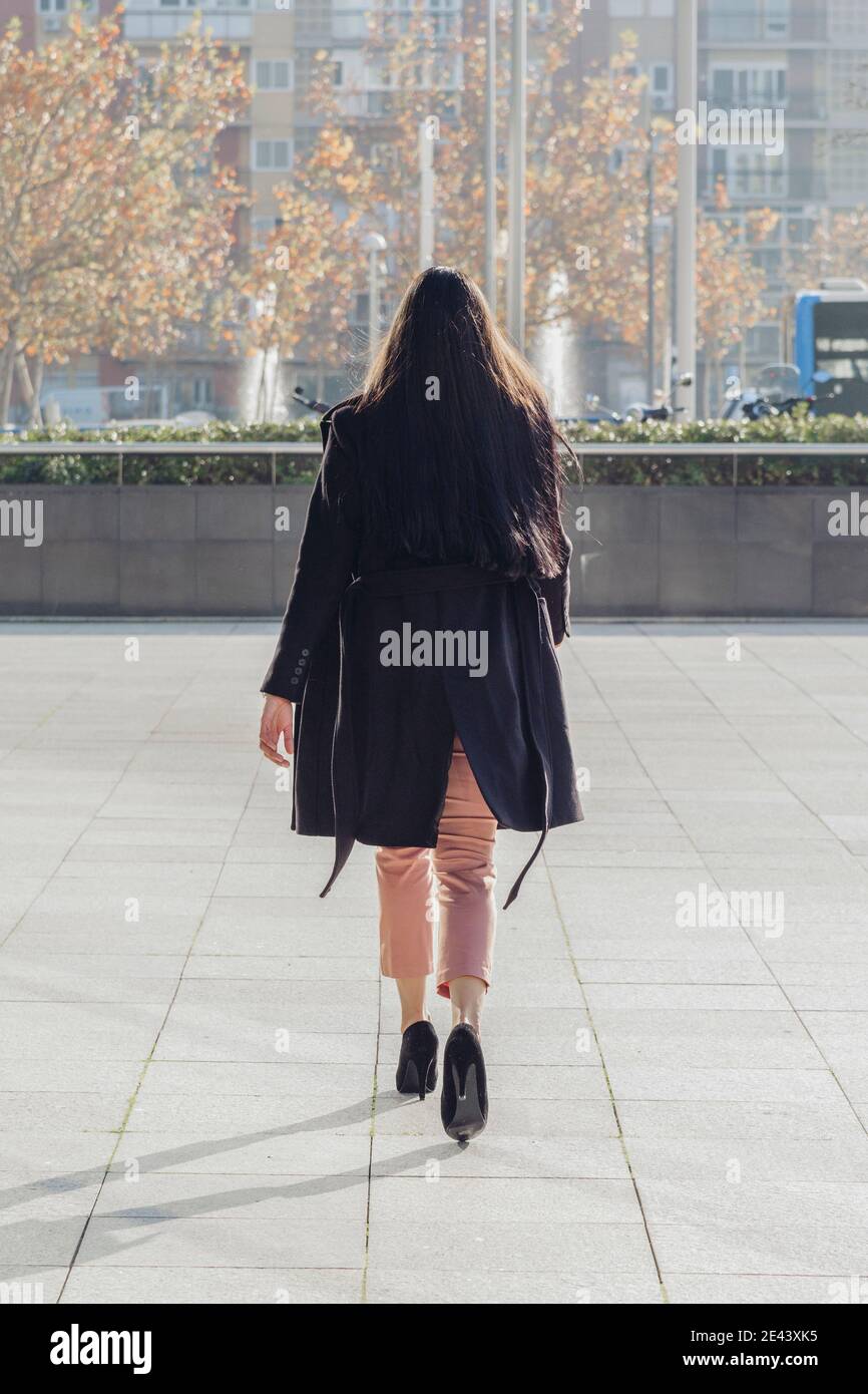 Vue arrière d'une jeune femme à poil long et élégante méconnaissable manteau  noir élégant et talons hauts marchant sur une place pavée en ville en  automne ensoleillé Photo Stock - Alamy