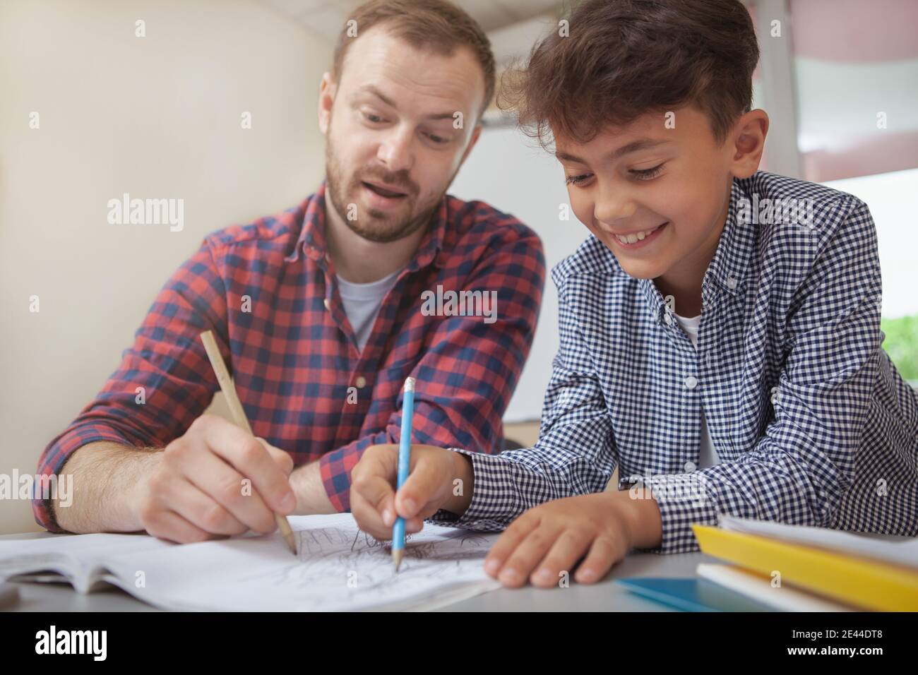 Plan court d'un charmant garçon d'école souriant joyeusement, en train de dessiner avec son professeur pendant la classe d'art. Enseignant aidant son petit étudiant avec l'artiste Banque D'Images