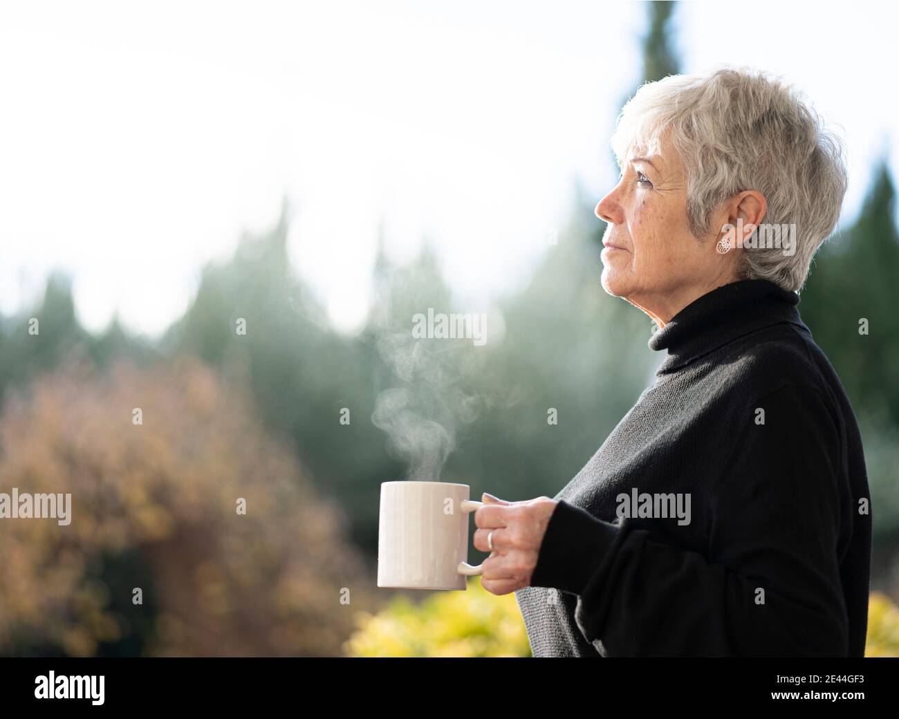 Femme âgée vêtue de cheveux noirs et blancs buvant une tasse de café près de la fenêtre à la maison en hiver Banque D'Images