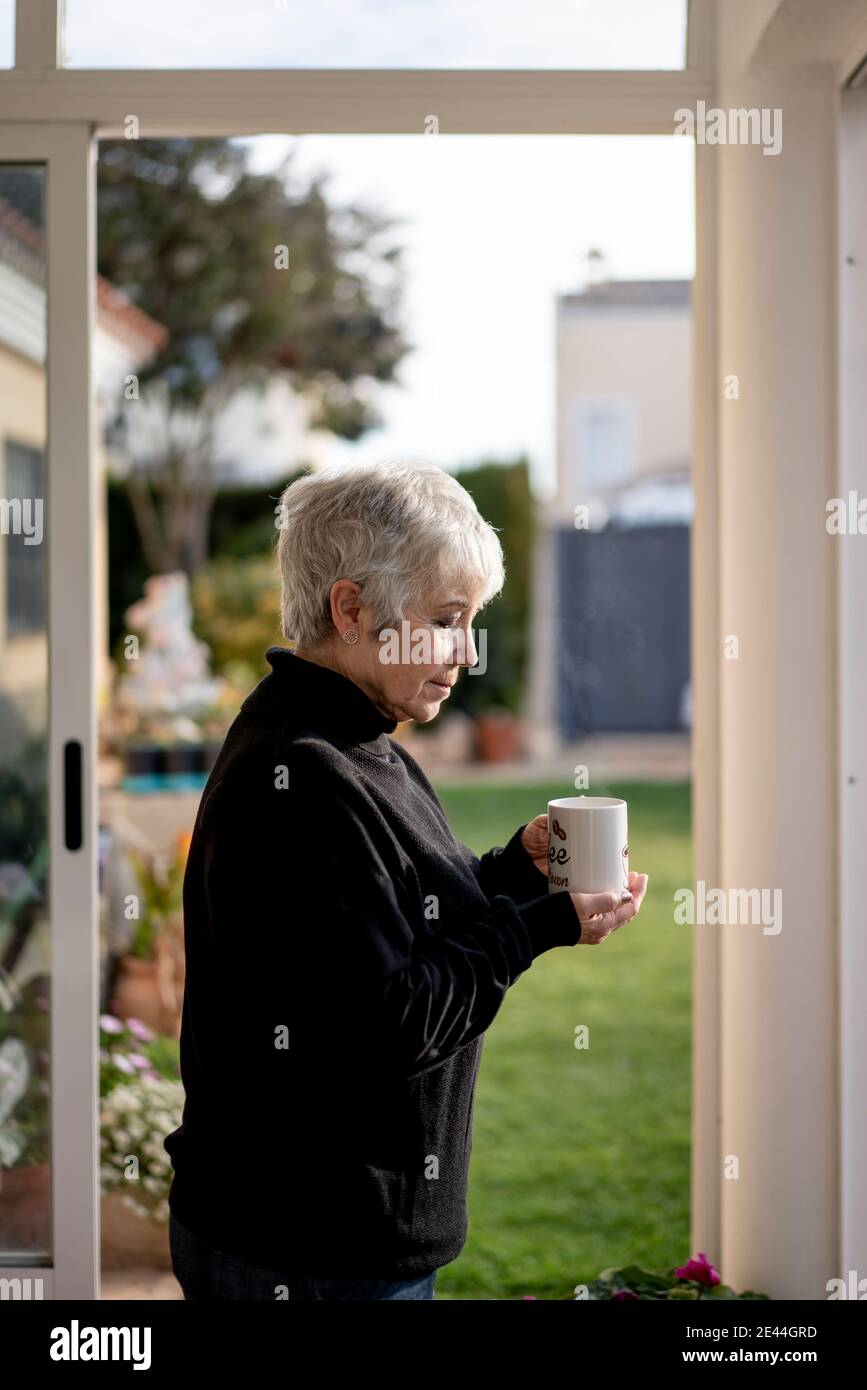 Femme âgée vêtue de cheveux noirs et blancs buvant une tasse de café près de la fenêtre à la maison en hiver Banque D'Images