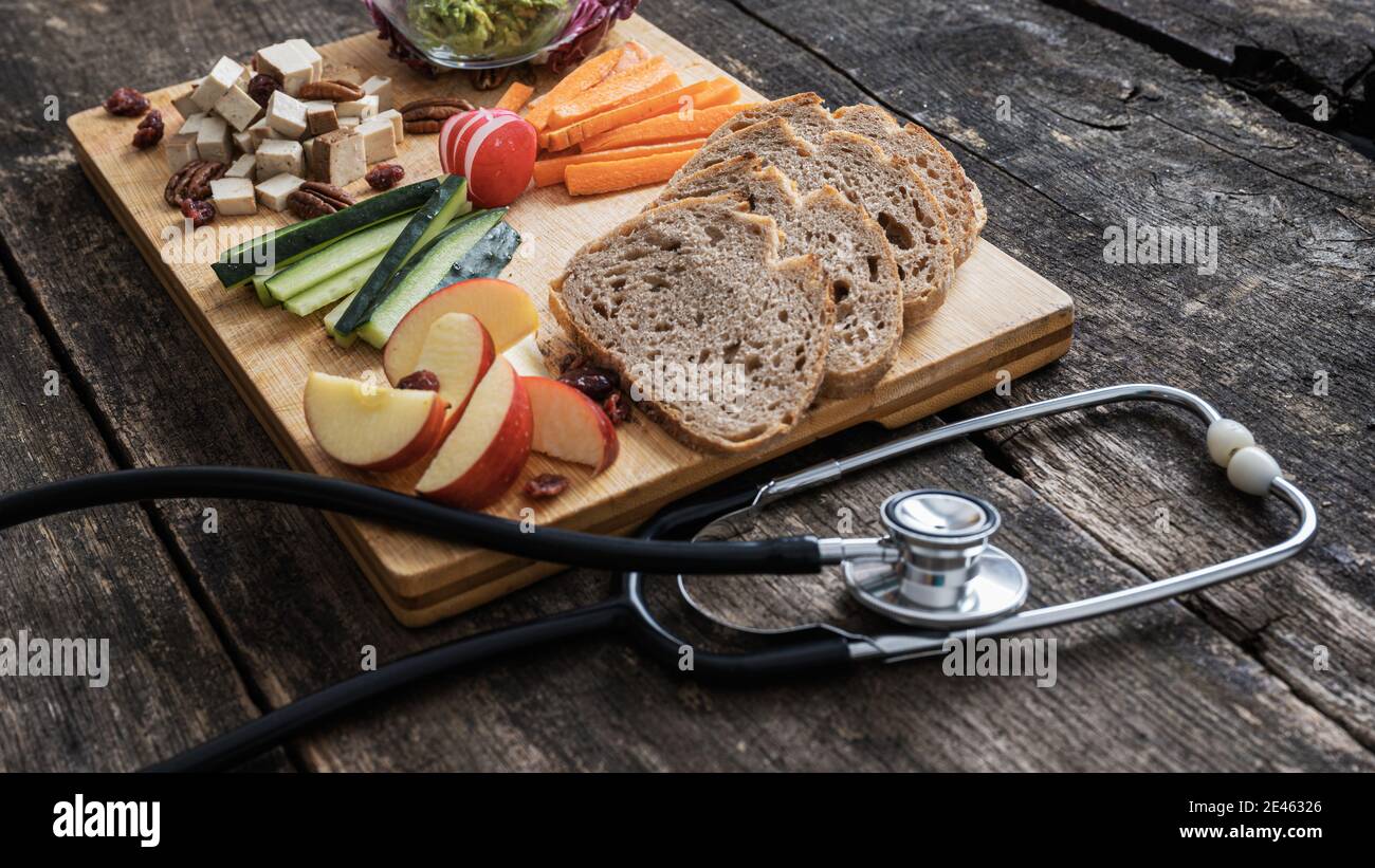 Image conceptuelle de régime sain - assiette de service en bois pleine de légumes, pain de levain de fruits et protéine végétalienne à côté d'un stéthoscope. Banque D'Images