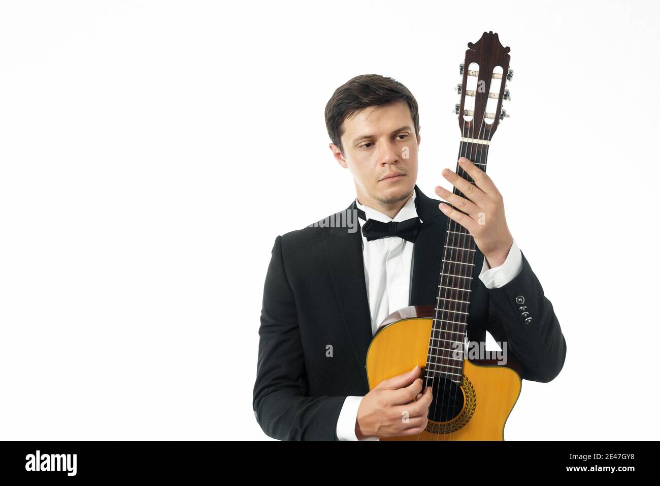 Jeune homme en costume classique noir posant avec une guitare classique et regardant le cou d'une guitare classique. En studio sur fond blanc Banque D'Images