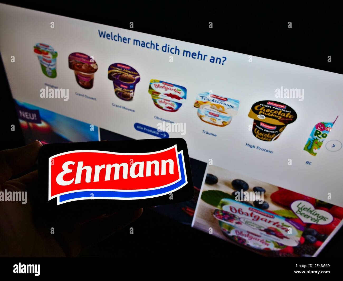 Personne tenant le téléphone mobile de l'entreprise laitière allemande Ehrmann AG avec le logo de la société sur l'affichage en face du site du produit. Mise au point sur l'écran du téléphone portable. Banque D'Images
