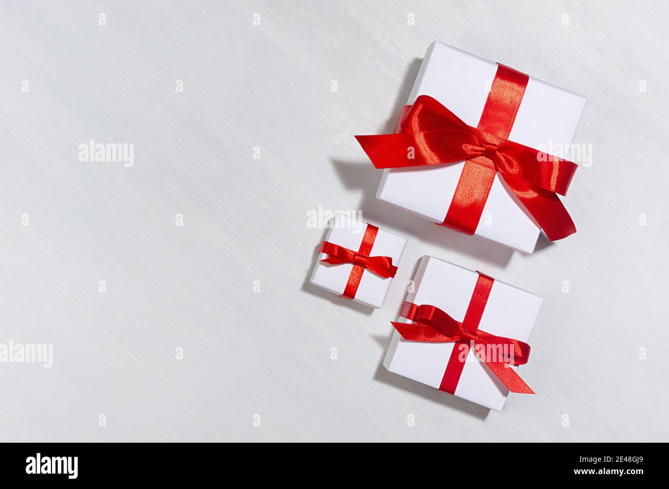Décor festif à vendre avec des boîtes-cadeaux blanches avec ruban de soie rouge au soleil avec ombre sur panneau de bois blanc, vue du dessus, bordure. Banque D'Images