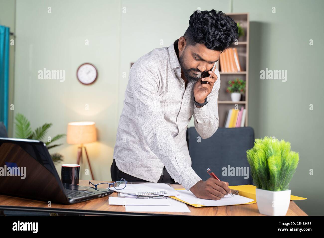 Jeune homme debout occupé à prendre des notes tout en parlant sur un téléphone mobile devant le bureau de travail - concept de l'appel d'affaires, parler avec les clients notant le faire Banque D'Images