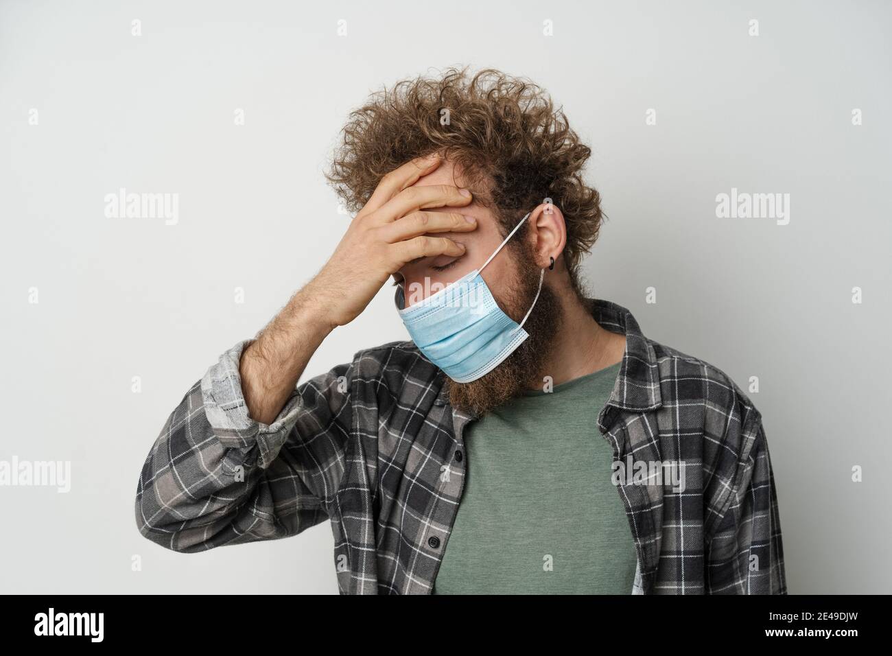 Souffrir de maux de tête portant un masque médical stérile de protection sur son visage pour protéger le coronavirus cheveux bouclés jeune homme portant du tissu écossais chemise et olive Banque D'Images