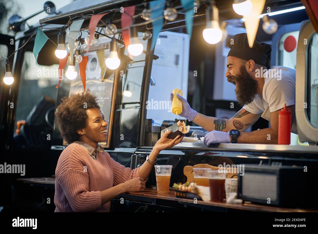 jeune employé de beardy caucasien dans la restauration rapide riant, ajoutant une moutarde dans un sandwich à une femme afro-américaine satisfait client Banque D'Images