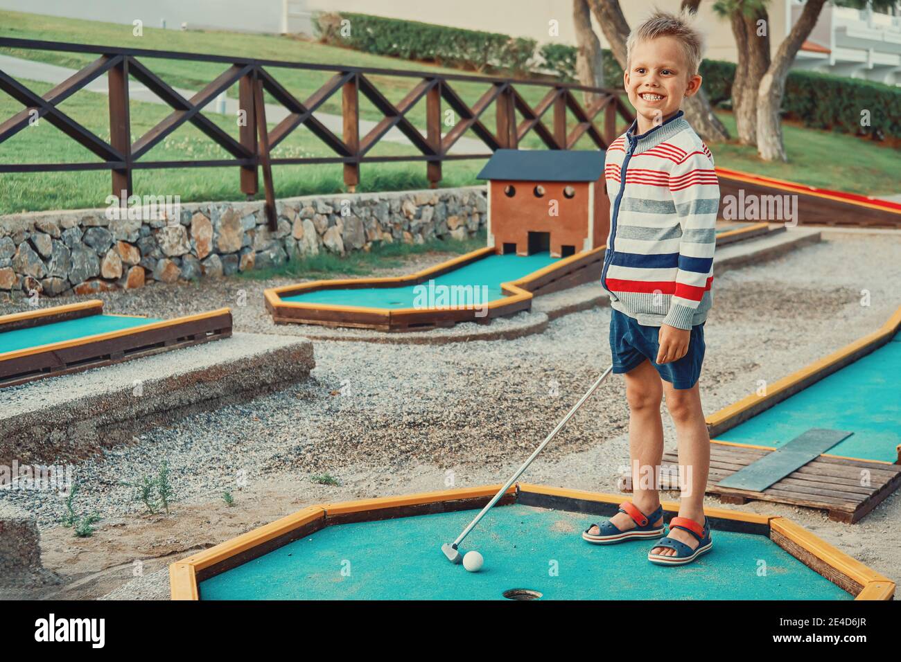 Joyeux garçon dans des vêtements décontractés souriant et regardant loin pendant jouer au mini-golf le jour d'été sur le territoire de l'hôtel Banque D'Images