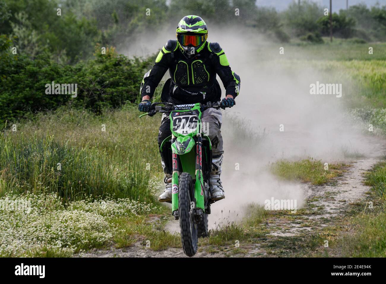 Un motocross de kawasaki conduit sur une route de terre surcultivée avec de l'herbe et des plantes et un nuage de poussière s'élève derrière lui. Banque D'Images