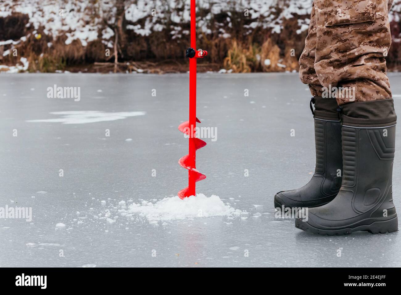 Pêcheur sans visage forant un trou avec une vis de glace pour la pêche sur glace. Pêche d'hiver. Homme jambes dans des bottes et glace vis sur fond de glace Banque D'Images