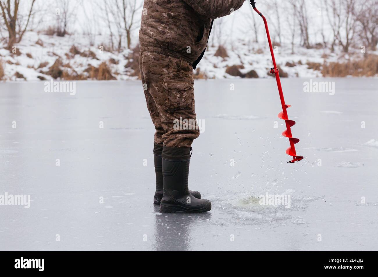 Pêcheur sans visage faisant le trou dans la glace neigeuse au lac par la vis de glace rouge. Concept de pêche d'hiver. Pêche sous la glace Banque D'Images