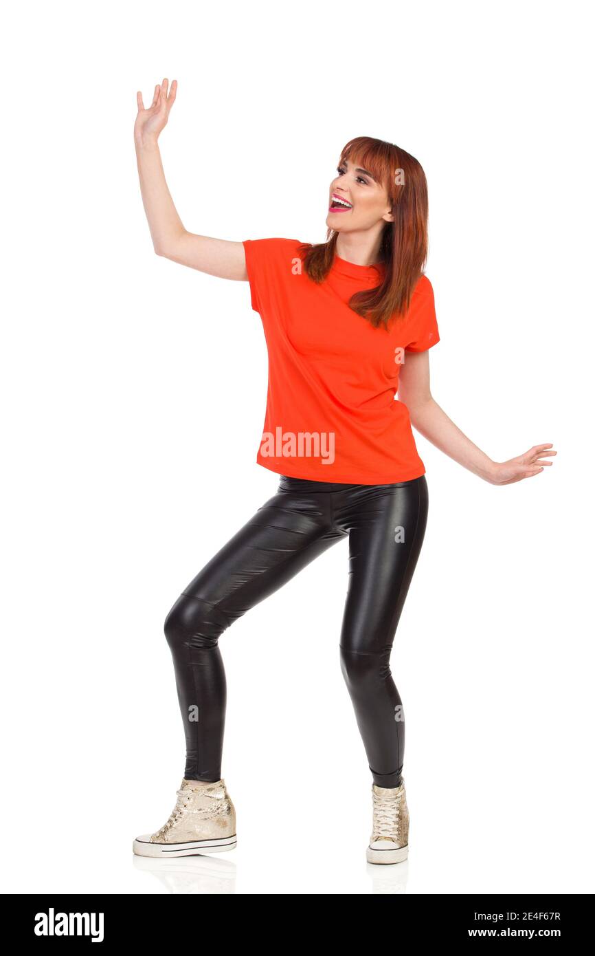 Une jeune femme en t-shirt orange, pantalon en cuir noir et baskets dorées danse, regarde loin et crie. Prise de vue en studio sur toute la longueur isolée sur blanc. Banque D'Images