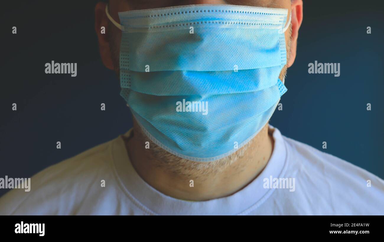Un homme à demi-visage portant un masque bleu médical pendant le verrouillage de l'épidémie de virus pose sur le mur bleu. Banque D'Images