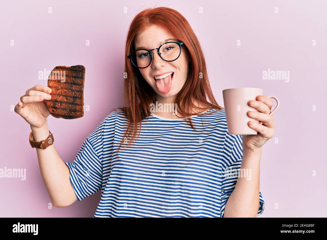 Jeune fille à tête rouge tenant des toasts brûlés pour le petit déjeuner collant la langue dehors heureux avec l'expression drôle. Banque D'Images