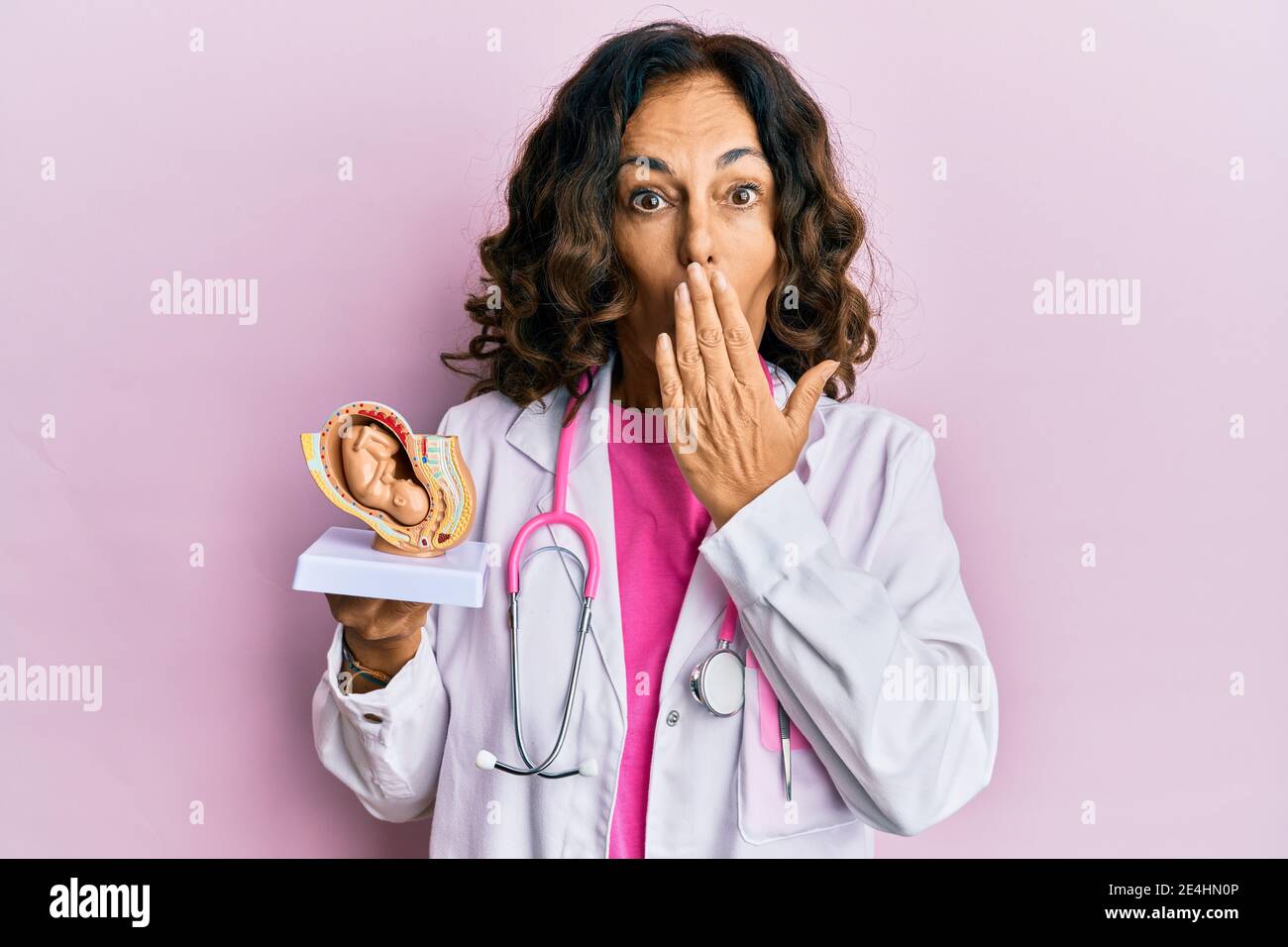 Moyenne âge hispanique médecin femme tenant le modèle anatomique de l'utérus femelle avec le fœtus couvrant la bouche avec la main, choqué et peur pour erreur. Surpri Banque D'Images