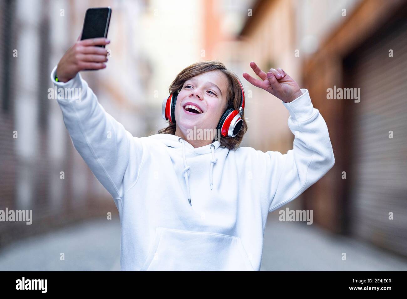 Un garçon souriant portant un casque pour un geste de paix tout en prenant un selfie par téléphone portable à l'extérieur Banque D'Images