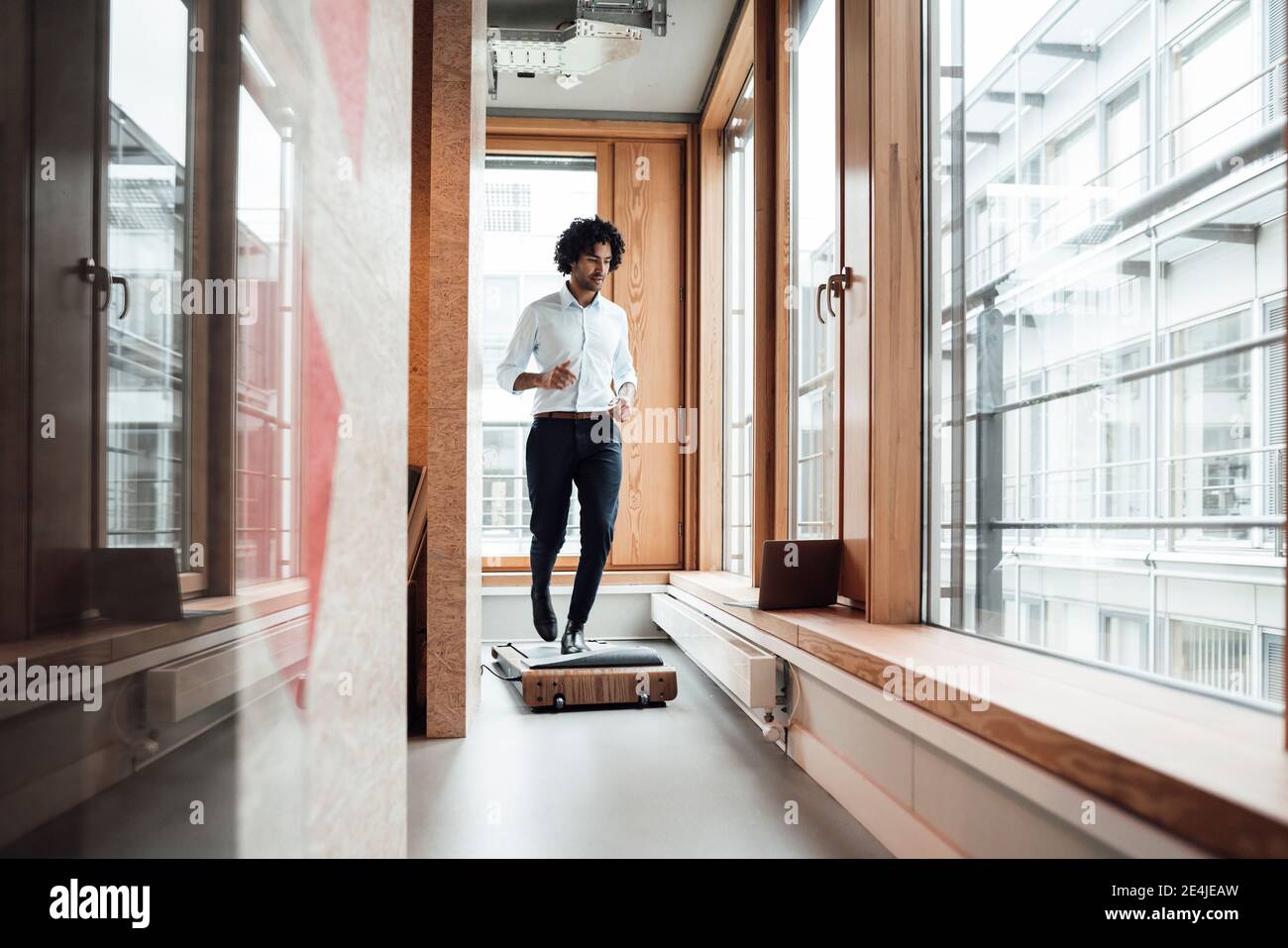 Homme d'affaires actif marchant sur un tapis roulant tout en regardant l'ordinateur portable contre la fenêtre dans le bureau Banque D'Images