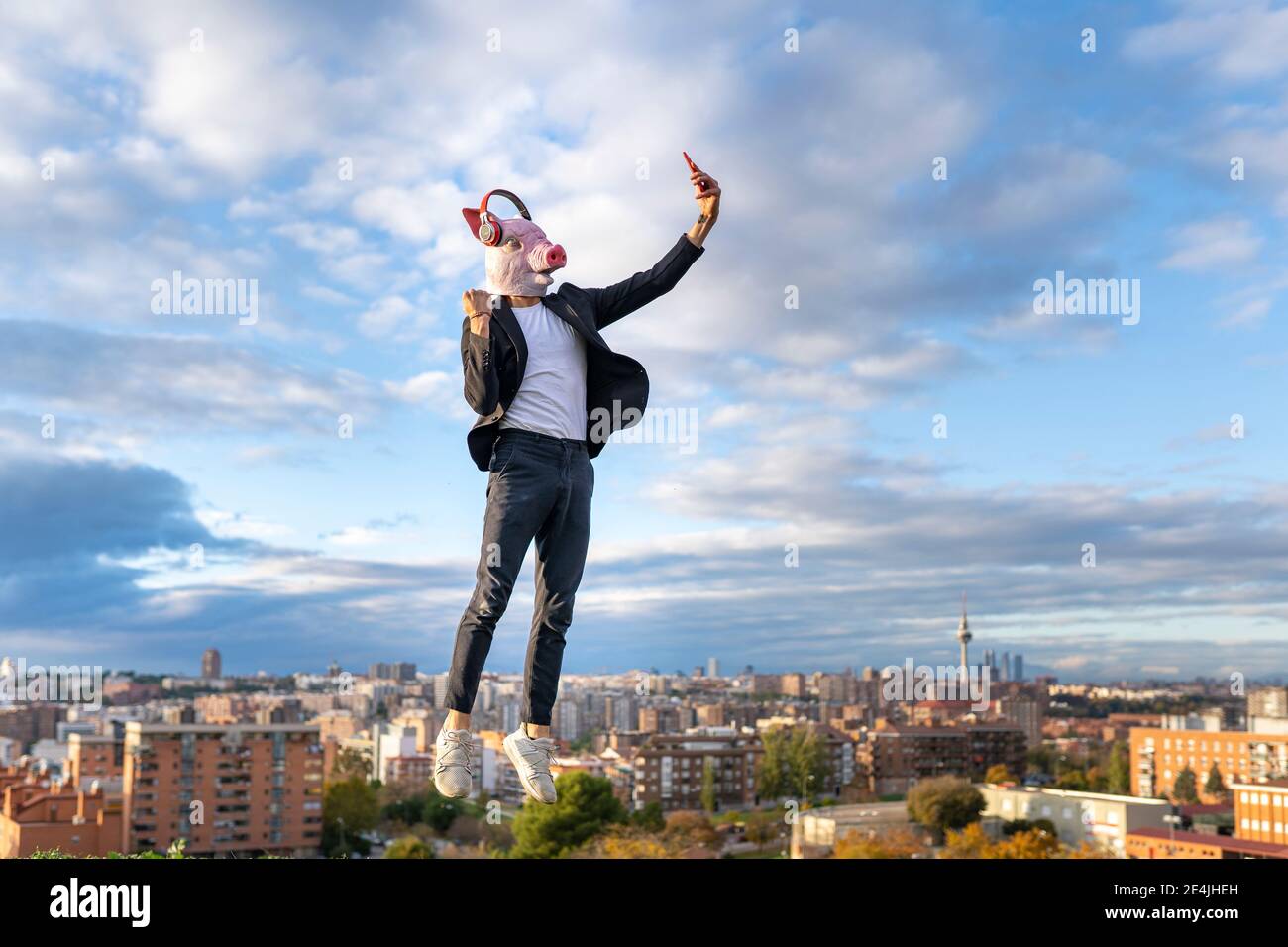 Homme d'affaires portant un masque de porc prenant selfie de smartphone pendant saut en ville contre le ciel Banque D'Images