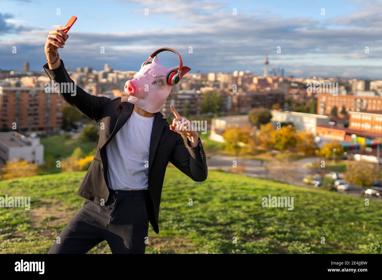 Homme d'affaires portant un masque de porc et un casque sans fil tout en prenant un selfie depuis un smartphone en pente Banque D'Images