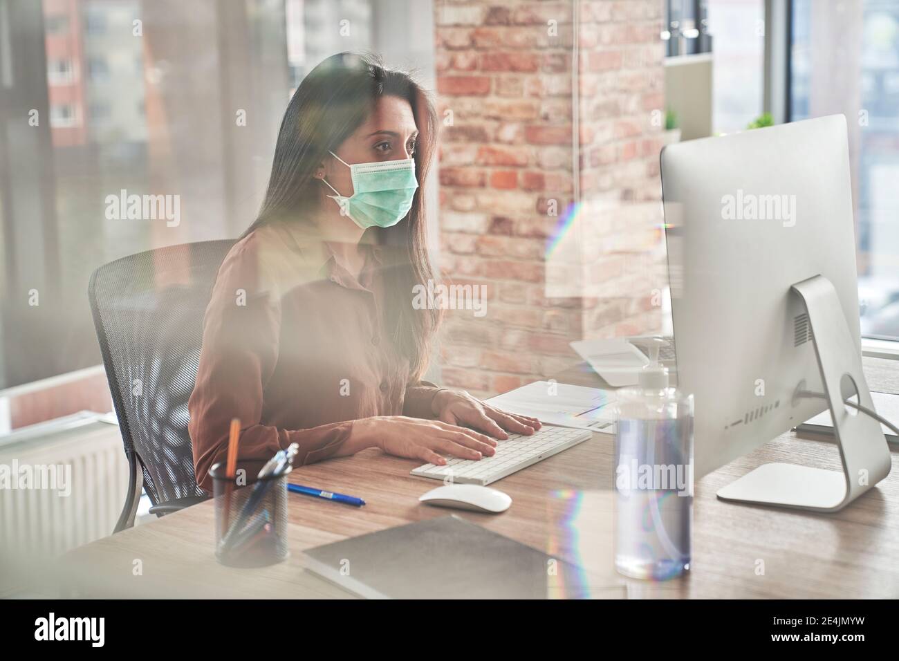 Femme adulte de taille moyenne travaillant sur un ordinateur de bureau vu à travers le verre pendant une pandémie Banque D'Images