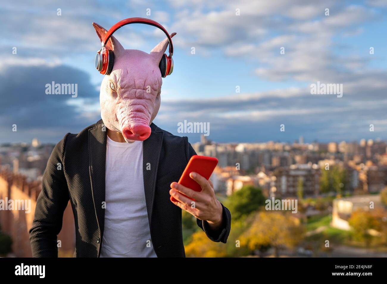 Homme d'affaires portant un masque de porc et un casque sans fil utilisant un smartphone contre le ciel Banque D'Images