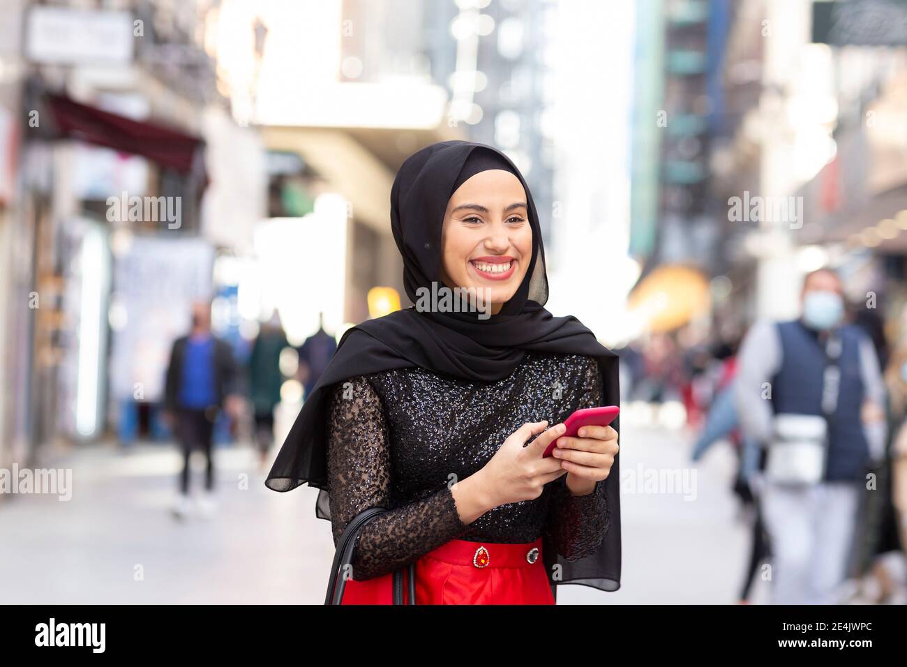 Portrait de la jeune belle femme portant le hijab noir souriant sur le trottoir avec smartphone entre vos mains Banque D'Images