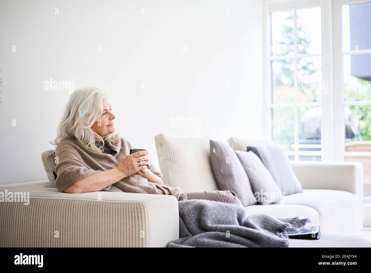 Femme attentionnés avec de longs cheveux blancs tenant une tasse de café pendant salon sur un canapé dans l'appartement Banque D'Images