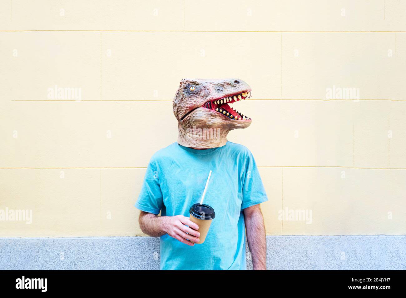Homme portant un masque de dinosaure avec un café debout contre le mur en ville Banque D'Images