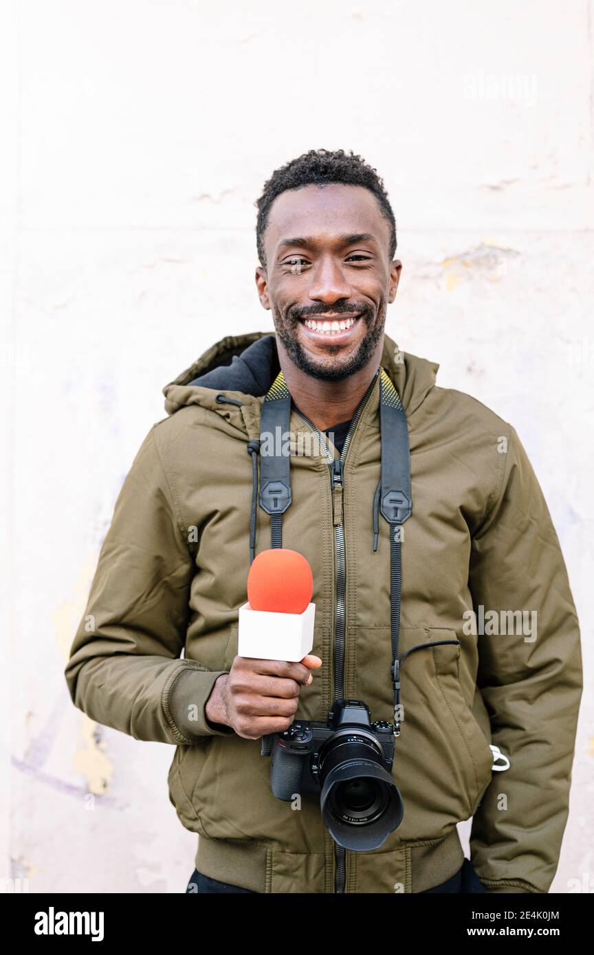 Journaliste masculin souriant avec microphone et caméra debout contre le blanc mur Banque D'Images