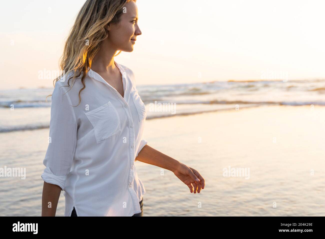 Femme souriante regardant loin en se tenant contre la mer pendant le coucher du soleil Banque D'Images
