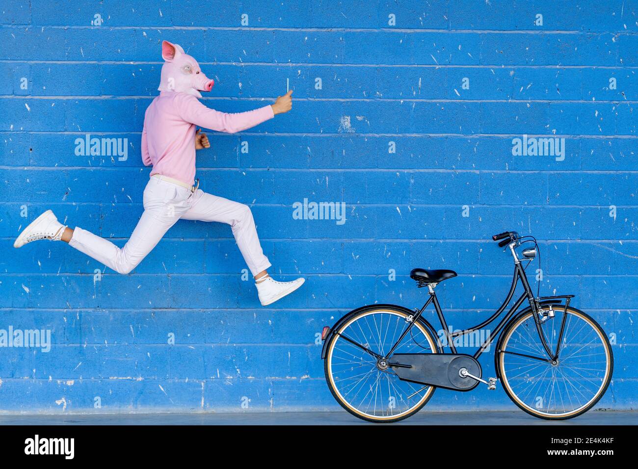 Jeune homme avec masque de porc sur le visage prenant selfie pendant saut contre le mur bleu Banque D'Images