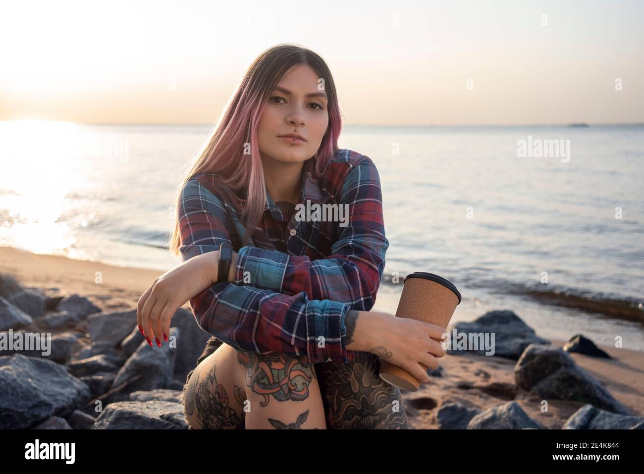 Jeune femme tendance avec tatouage tenant une tasse de café réutilisable contre mer à la plage Banque D'Images