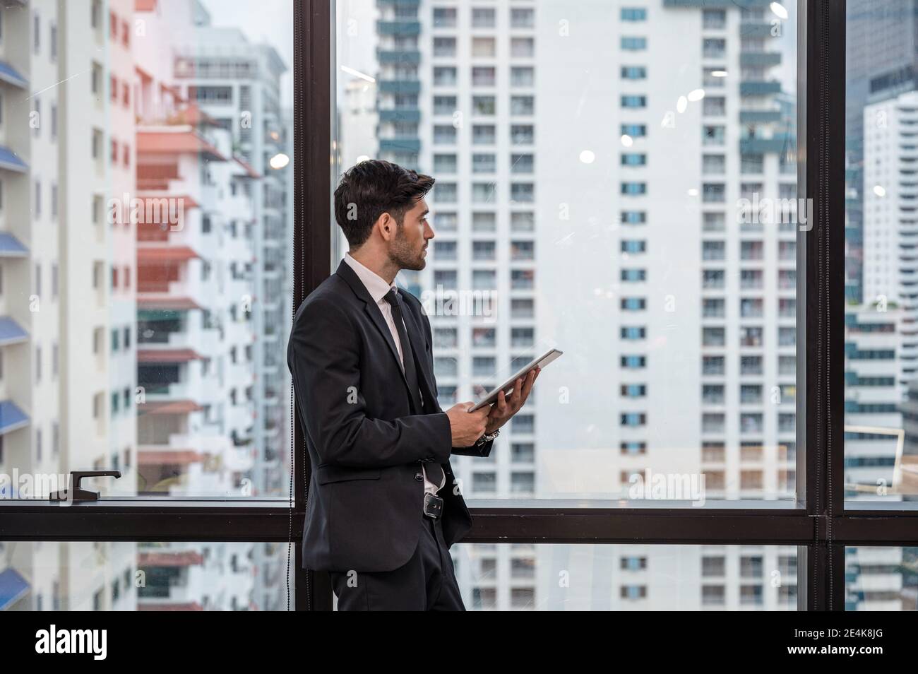 Homme d'affaires visionnaire caucasien en costume debout avec tenir tablette et en regardant par la fenêtre dans le bureau Banque D'Images