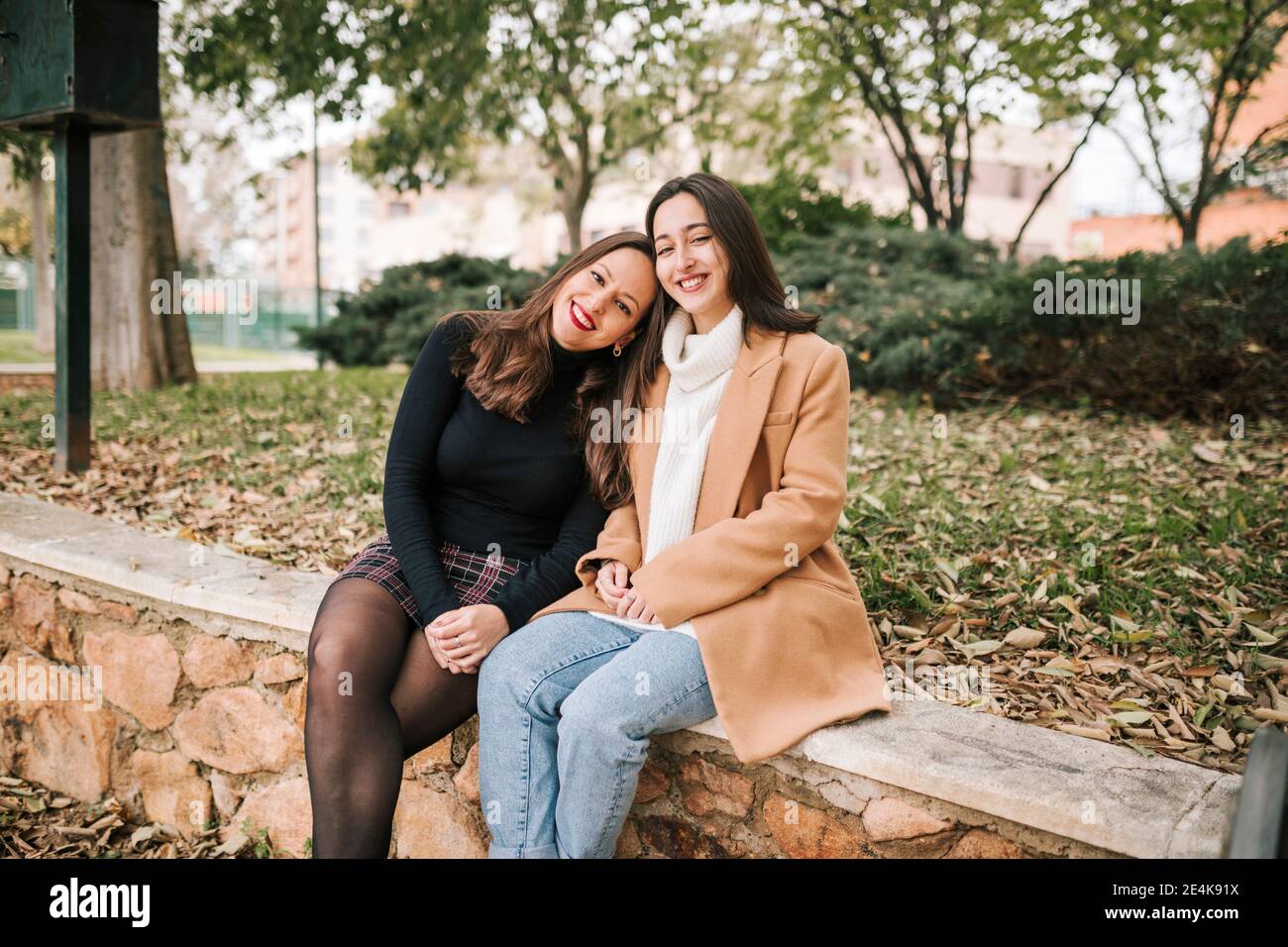 Mère souriante avec la tête sur l'épaule de la fille assise sur retenue mur dans le parc Banque D'Images