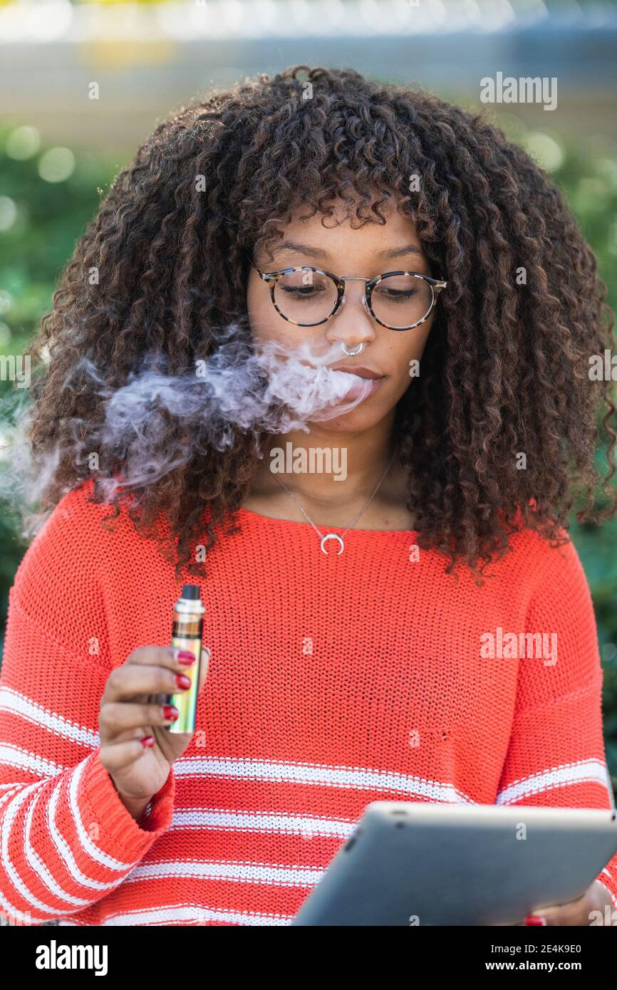 Femme afro portant des lunettes fumant la cigarette électronique tout en utilisant le numérique tablette au parc Banque D'Images