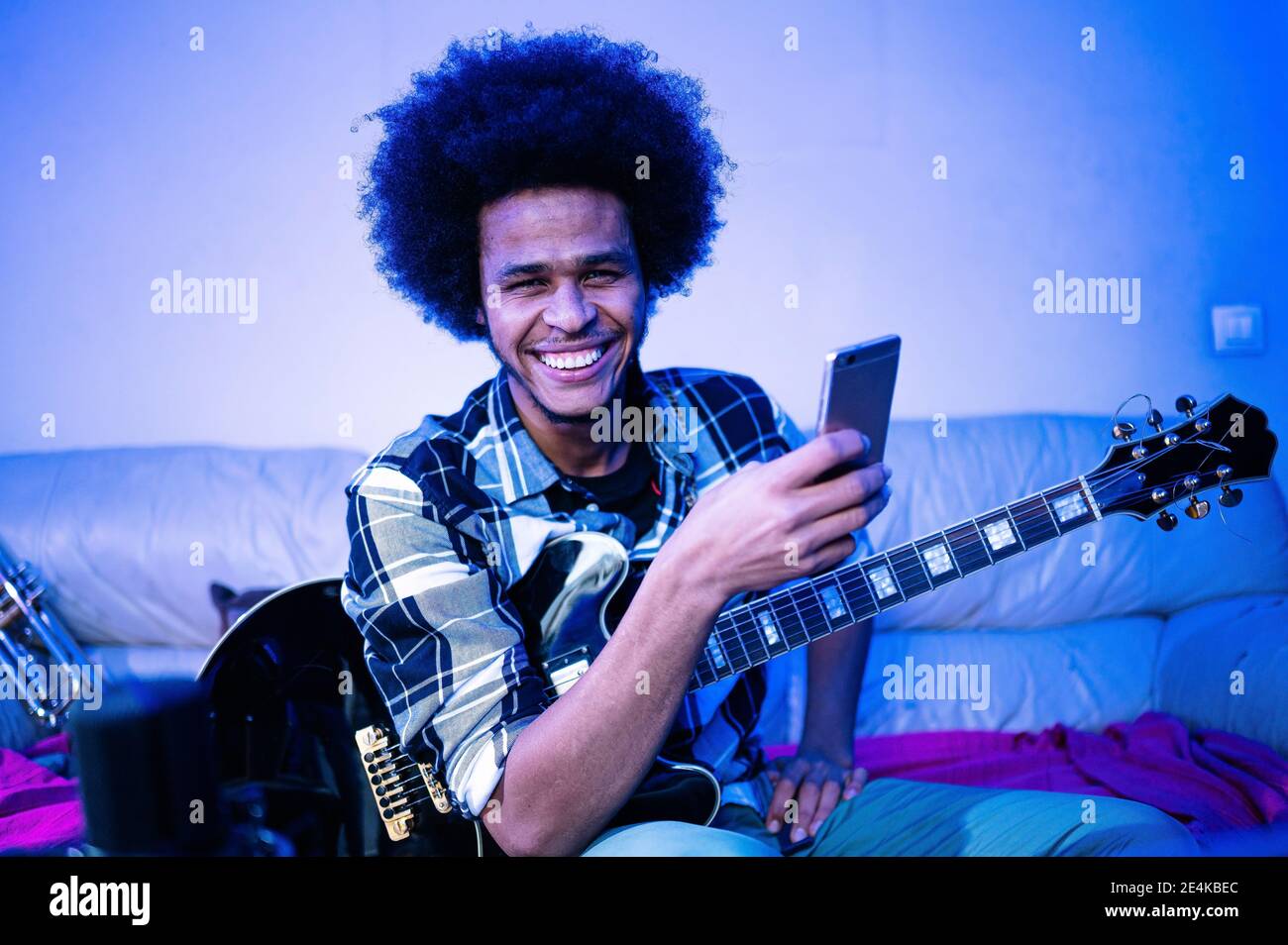 Joyeux afro homme avec la guitare regardant loin tout en tenant le téléphone mobile dans la pièce bleue Banque D'Images