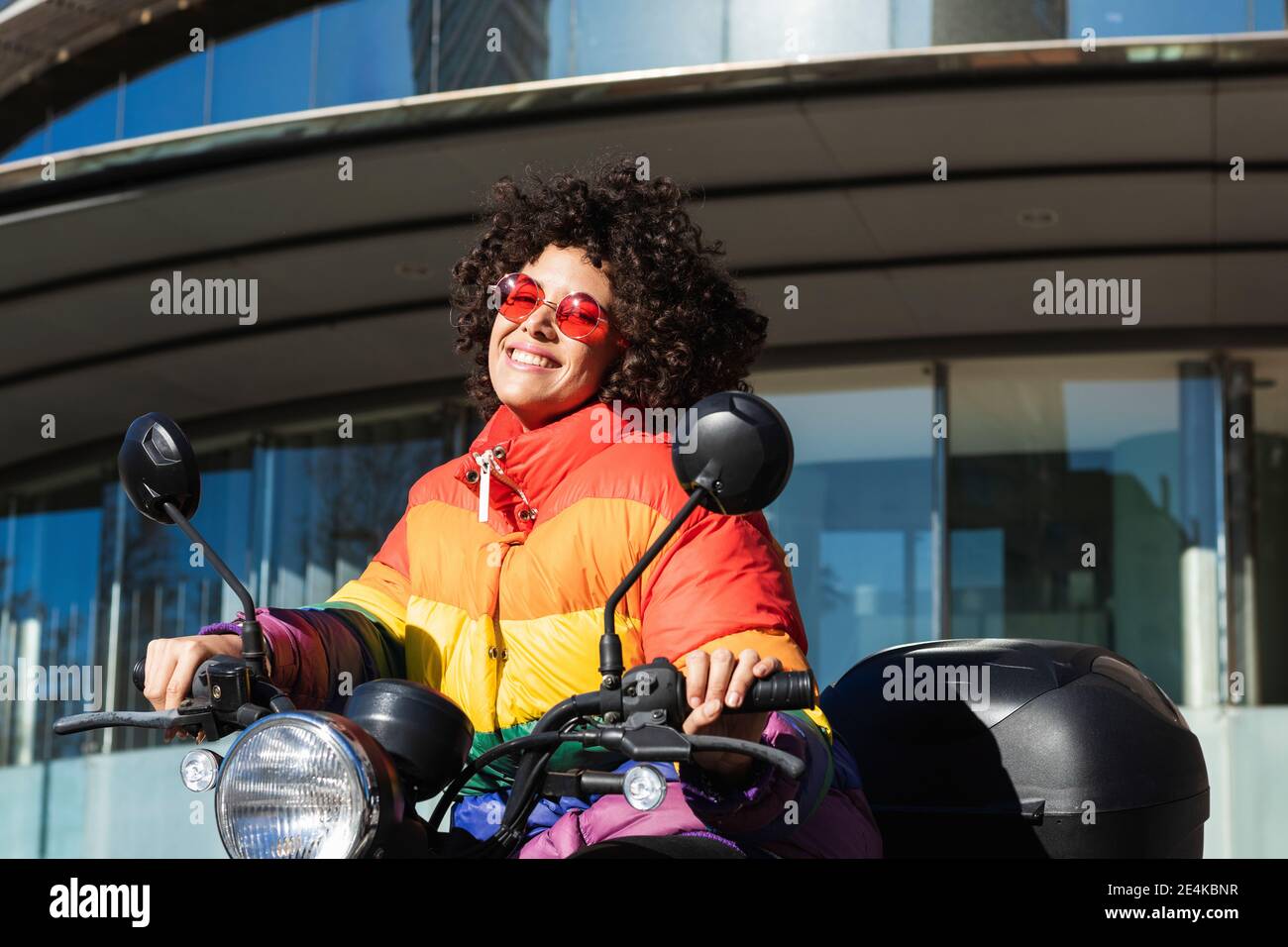 Femme portant des lunettes de soleil et une veste colorée souriant lorsqu'elle est assise moto Banque D'Images