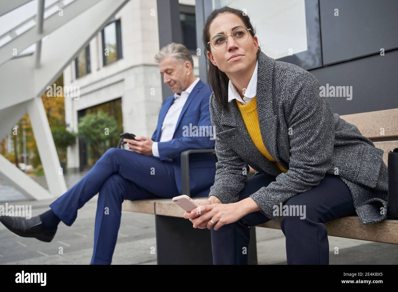 Navetteurs hommes et femmes avec smartphones assis sur un banc en attendant à l'arrêt de bus Banque D'Images