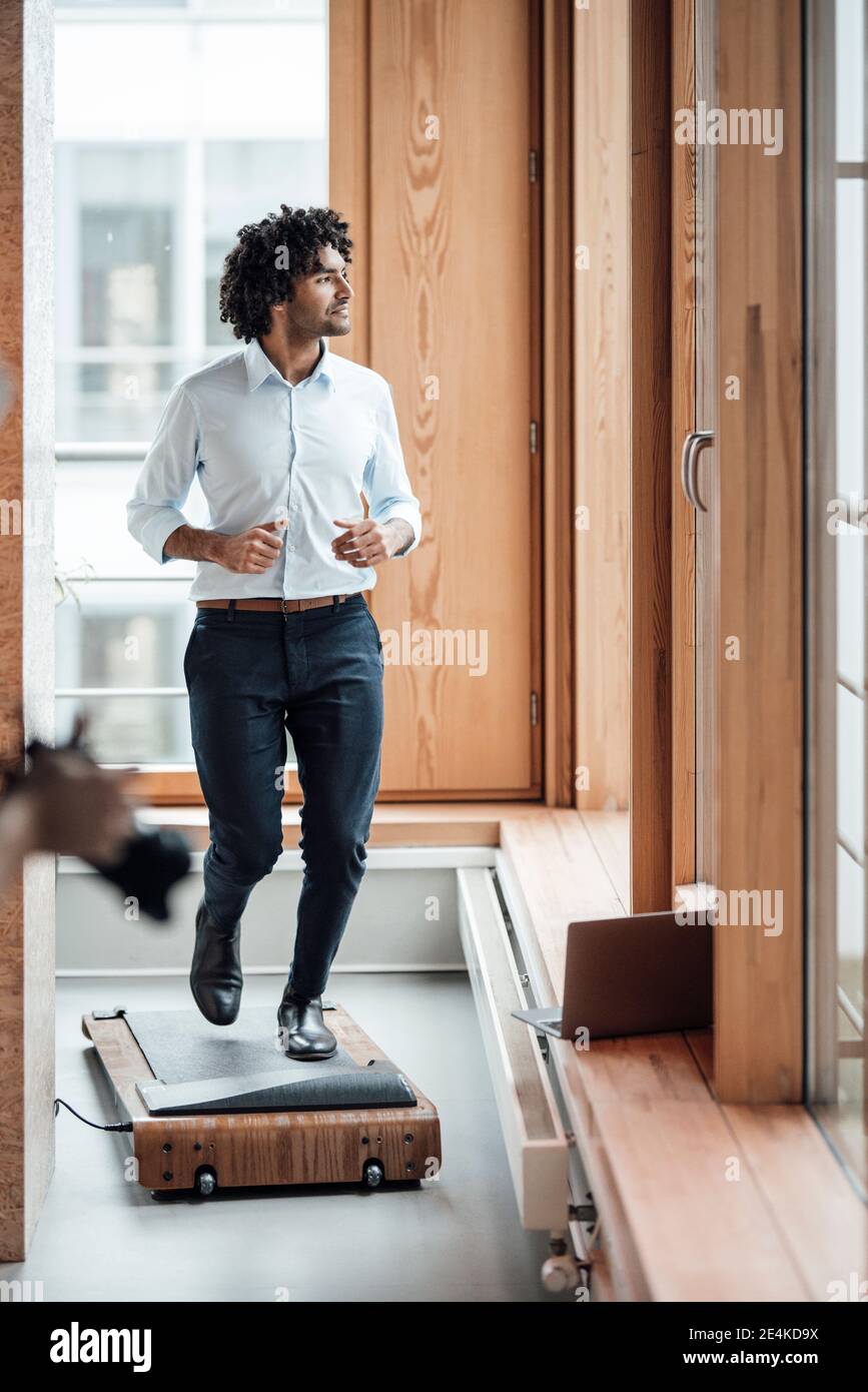 Un homme actif qui fait du jogging sur un tapis roulant tout en regardant contre fenêtre sur le poste de travail Banque D'Images